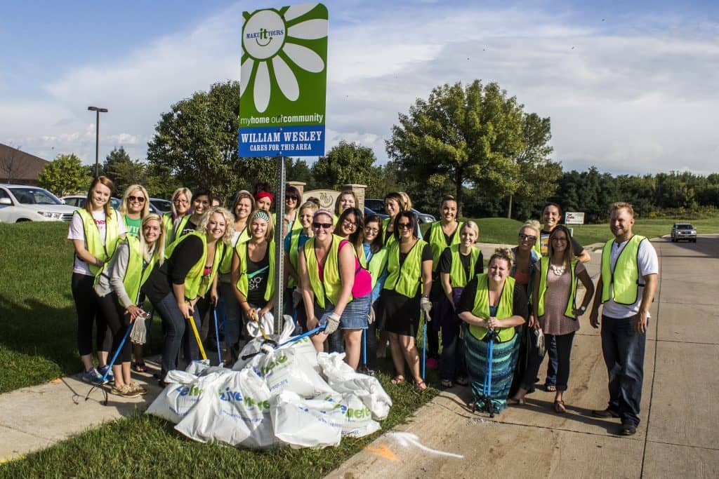 Volunteers wearing yellow high vis vests stand with litter tongs and trash bags near MAKE it YOURS adoption sign.